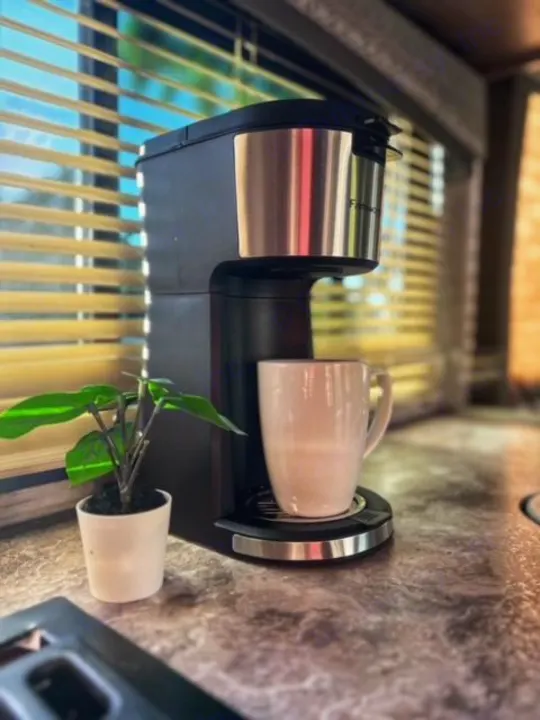 An RV rental kitchen features a black coffee maker with a white mug on a speckled counter. Sunlit blinds are visible in the background.. Thor Four Winds 2022