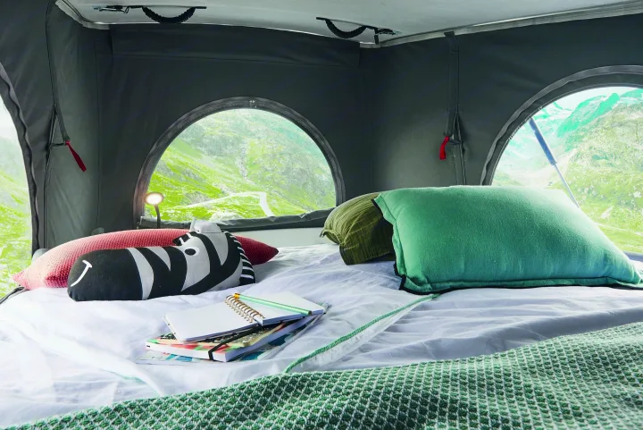 Cozy RV rental bed with a stunning mountain view from arched windows. Features pillows, a blanket, and notebooks for planning adventures.. Pilote V600 Toit relevable 2025