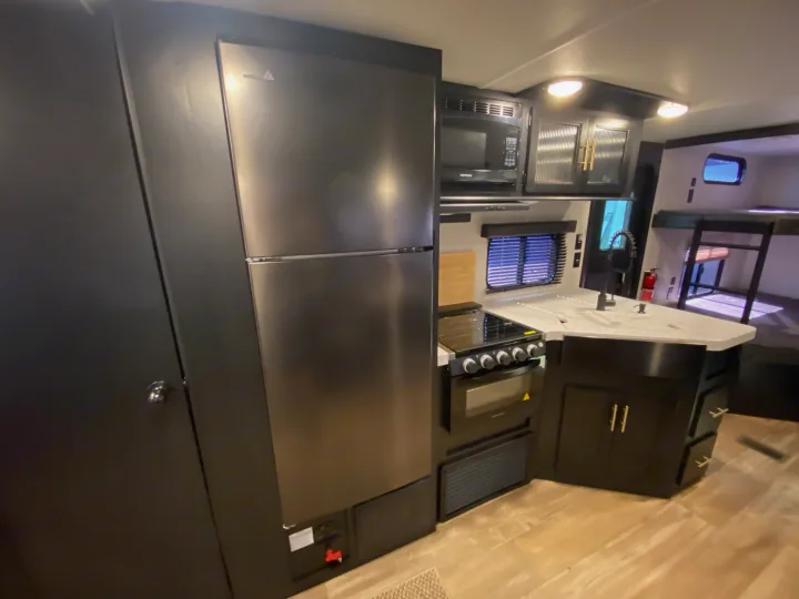 Modern RV rental kitchen with stainless steel fridge, microwave, oven, and sink on light counters. Bunk beds visible in background.. Forest River Cherokee 2021