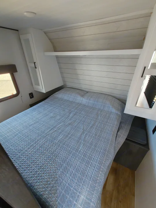 RV rental bedroom: Features a blue patterned bed, white overhead cabinets, wood-look walls, and a side window.. Keystone RV Hideout 2023