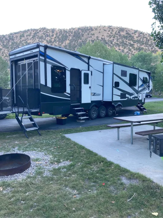 Spacious RV rental set up at a tranquil mountain campsite, featuring a picnic table, fire pit, and grill. Ideal outdoor escape.. Keystone Fuzion 2021