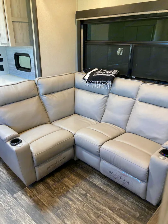 Modern RV rental interior: L-shaped sectional sofa with recliners, cup holders, and wood-look flooring. A window and kitchen area are visible.. Keystone Fuzion 2021