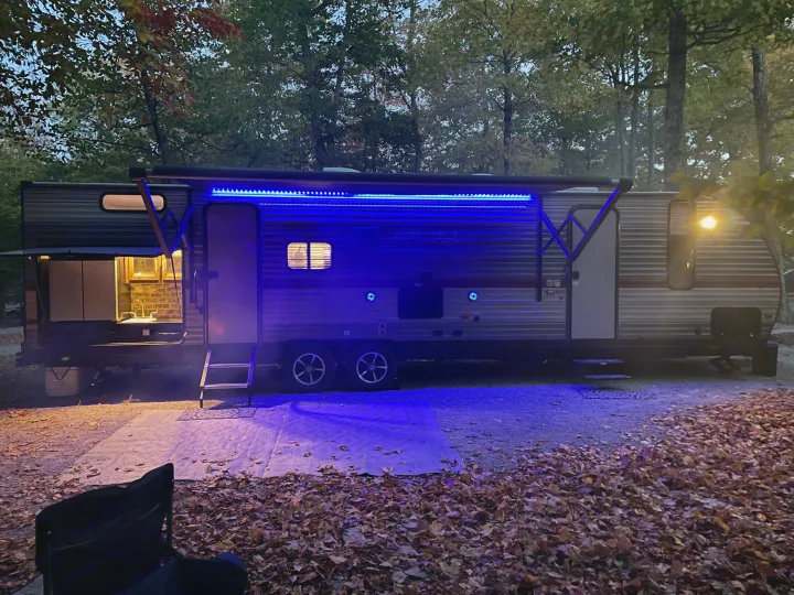 Nighttime view of an RV rental at a cozy campsite, with glowing blue awning lights and an outdoor kitchen. Ideal for fall camping.. Forest River Cherokee Grey Wolf 2019