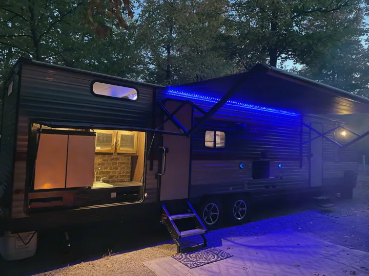 Evening RV rental: travel trailer with illuminated outdoor kitchen and blue LED awning lights at campsite.. Forest River Cherokee Grey Wolf 2019