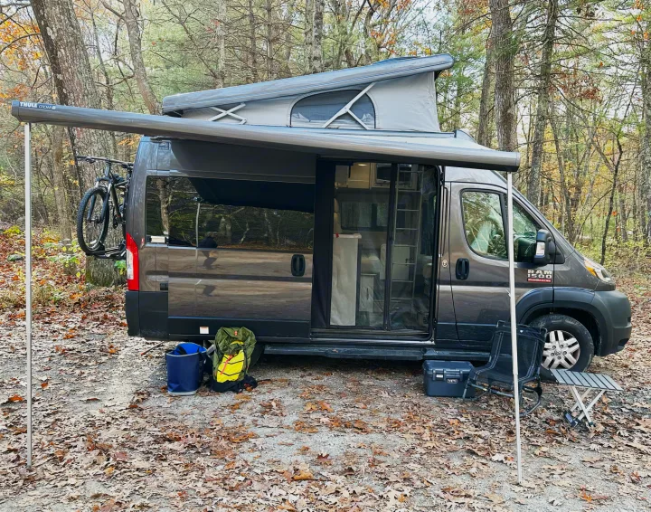 A grey pop-top RV rental with an extended Thule awning is set up in a leafy forest. A bike, chair, and table are visible.. Thor Motor Coach Rize 2022