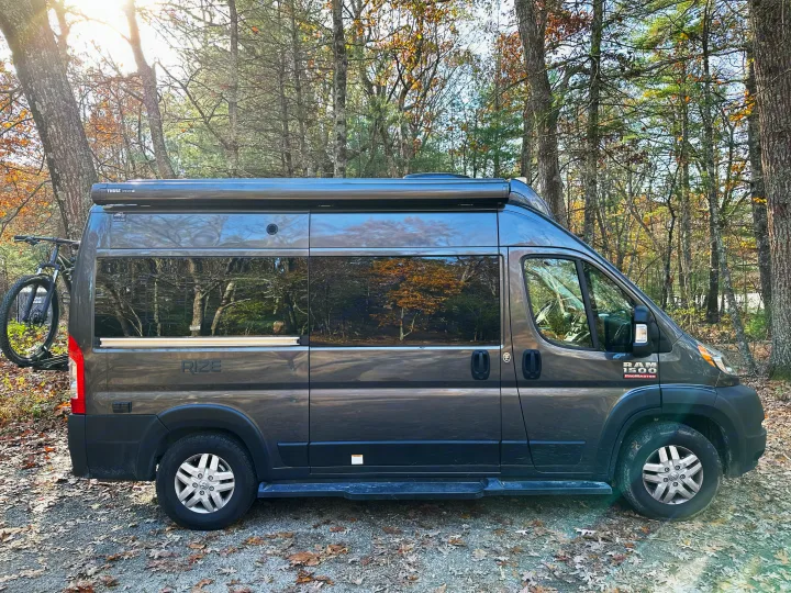 Explore nature with this grey Ram ProMaster camper van rv rental, featuring a bike rack, parked among sunlit trees.. Thor Motor Coach Rize 2022