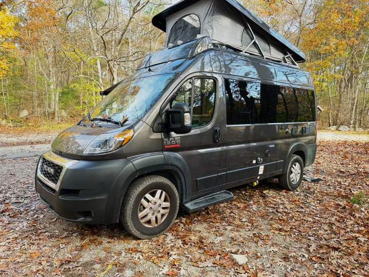 A grey Ram ProMaster camper van with a pop-top roof is parked on a leaf-strewn path in an autumn forest, ready for rv rental.. Thor Motor Coach Rize 2022