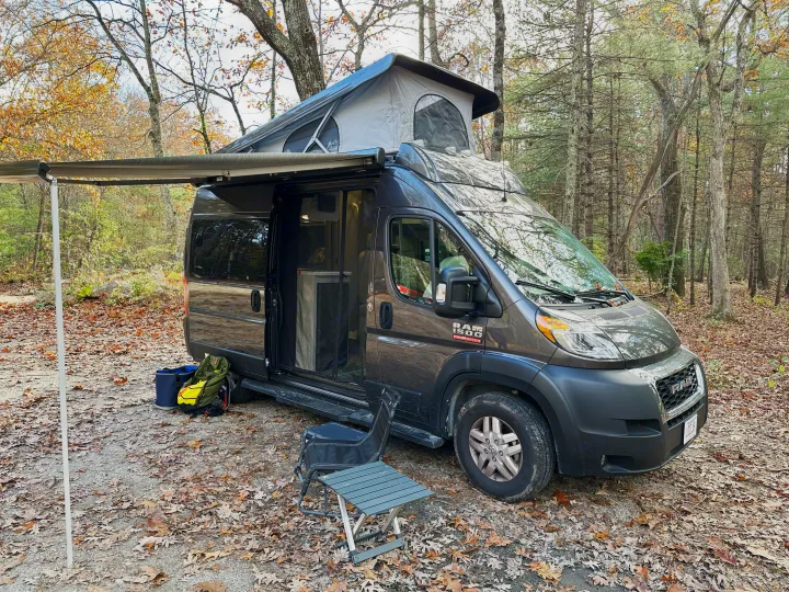 Grey Ram ProMaster RV rental with pop-top extended & awning out, parked in a scenic autumn forest campsite.. Thor Motor Coach Rize 2022