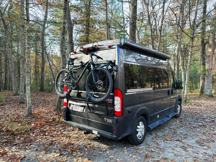 Dark grey RV rental, a camper van with two bikes on its rear rack, parked among trees with fallen autumn leaves.. Thor Motor Coach Rize 2022