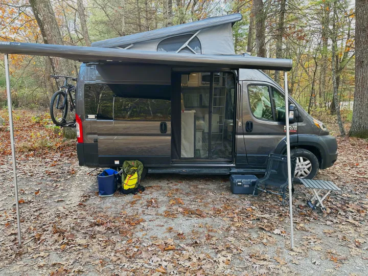 Dark grey camper van with pop-top roof and extended awning, perfect for an rv rental outdoor adventure. Bike, cooler, and camping gear seen.. Thor Motor Coach Rize 2022