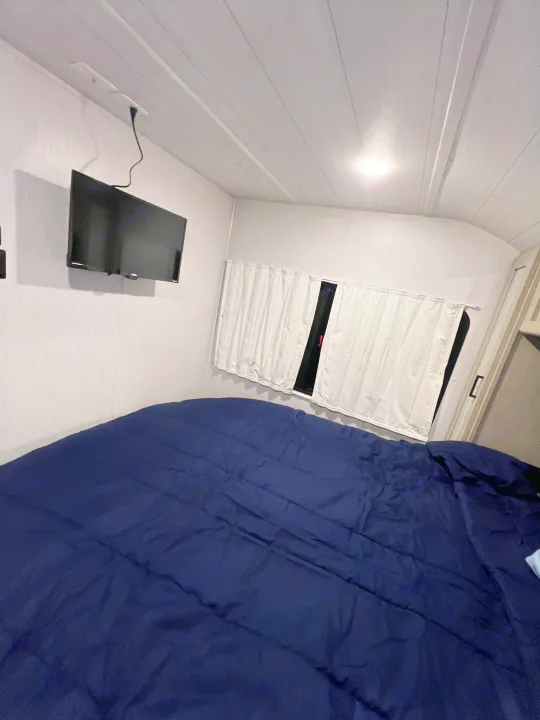 Inside a compact RV rental bedroom featuring a comfy navy bed, wall-mounted TV, and window coverings.. Forest River 31KQBTS 2024