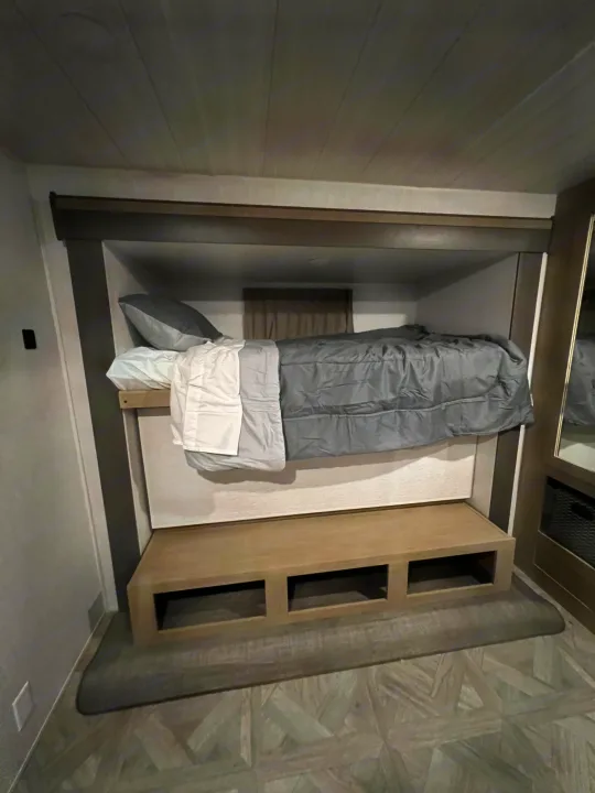 RV rental interior: A cozy bunk bed with grey and white linens sits above a functional storage bench.. Forest River 31KQBTS 2024