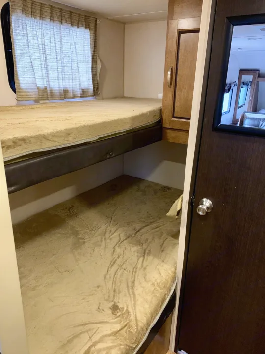 Compact bunk beds inside an RV rental, featuring light bedding, a window with curtains, and an adjacent cabinet and door.. Forest River Cruise Lite 2017