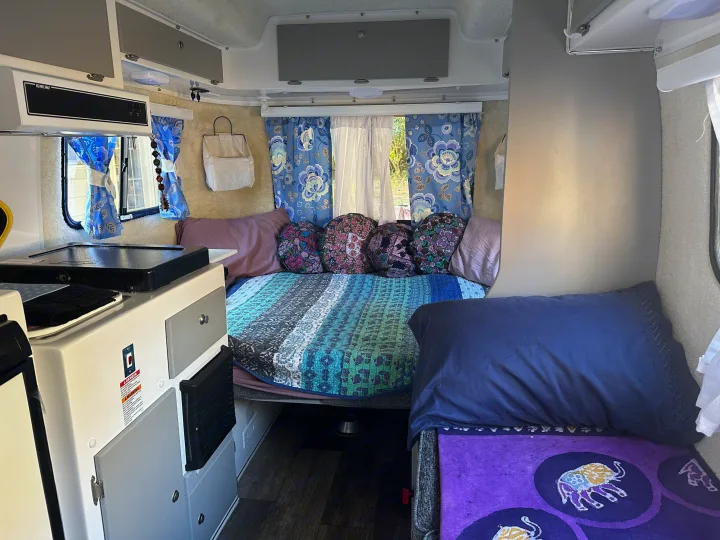 Inside a compact rv rental: a cozy bed with quilted bedding, blue curtains, and a small kitchen area with overhead storage.. Casita Spirit 2023