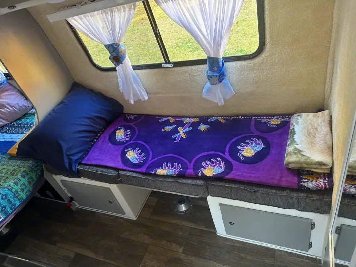 Cozy rv rental interior: A bed with a purple blanket featuring elephants and dragonflies, a blue pillow, and tie-back curtains.. Casita Spirit 2023