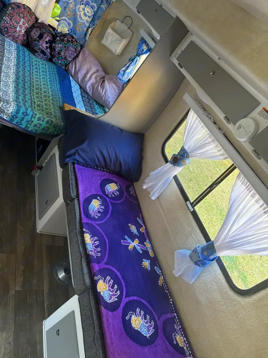 Cozy RV rental interior: colorful bedding, elephant throw on seating, sheer window curtains with green view, and smart storage.. Casita Spirit 2023