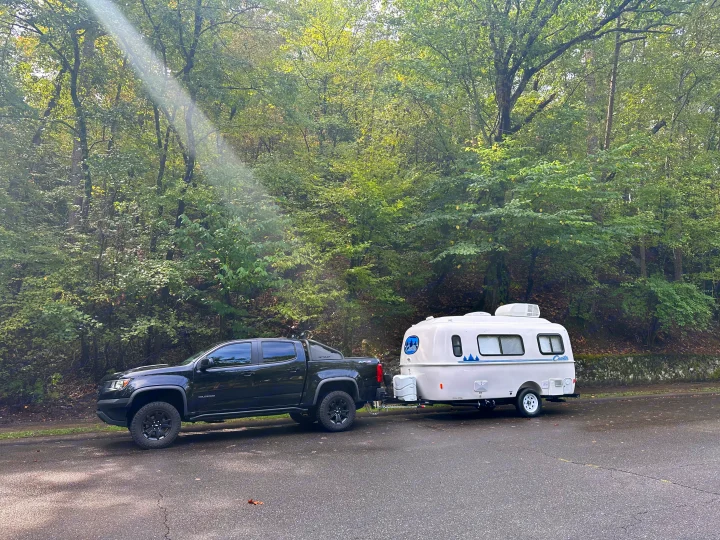 A black pickup truck is hitched to a white travel trailer on an asphalt road, backed by a lush forest. Consider this RV rental for camping.. Casita Spirit 2023