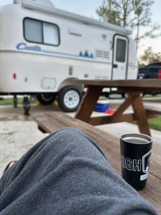 Relaxing at our RV rental campsite. A person's legs and a coffee mug are on a picnic table, with a white Casita RV in the background.. Casita Spirit 2023