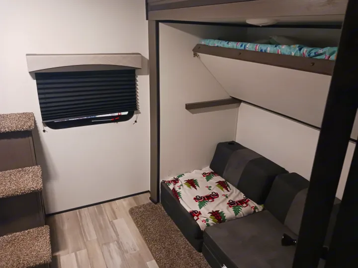 View inside an RV rental showing bunk beds (upper with blue bedding), a dark gray sofa, a window with pleated blind, and carpeted steps.. Embrace EL310 2021
