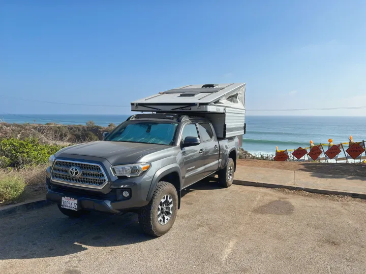 Scenic RV rental: Grey Toyota Tacoma truck with pop-up camper overlooks ocean waves, ideal for beach camping adventures.. Four Wheel Campers Fleet 2021