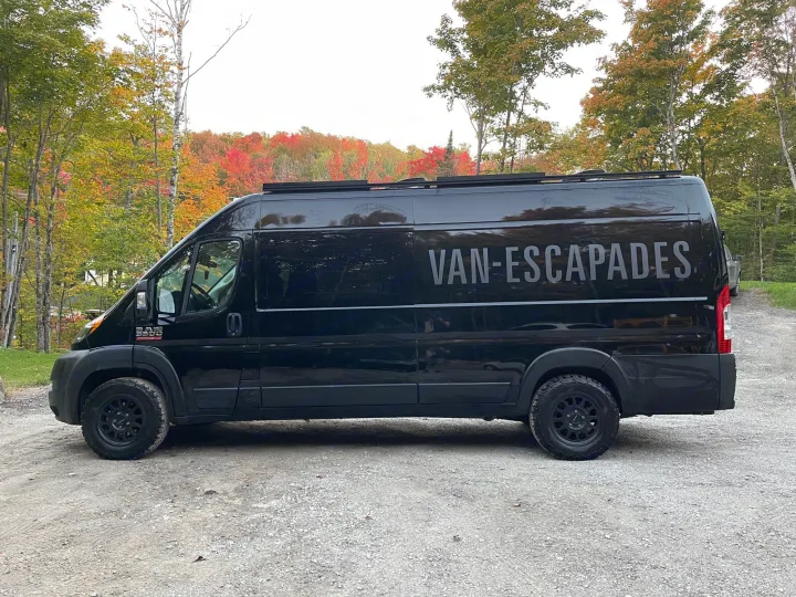 Explore nature with this black RV rental camper van parked on gravel, surrounded by vibrant fall trees.. RAM Promaster 2022