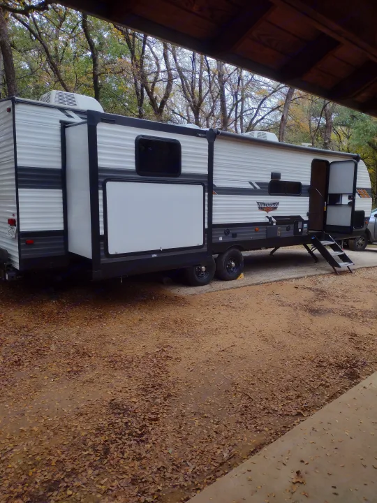 A white and grey travel trailer (RV rental) parked on a leaf-covered lot under autumn trees at a campground.. Forest River Wildwood 2022