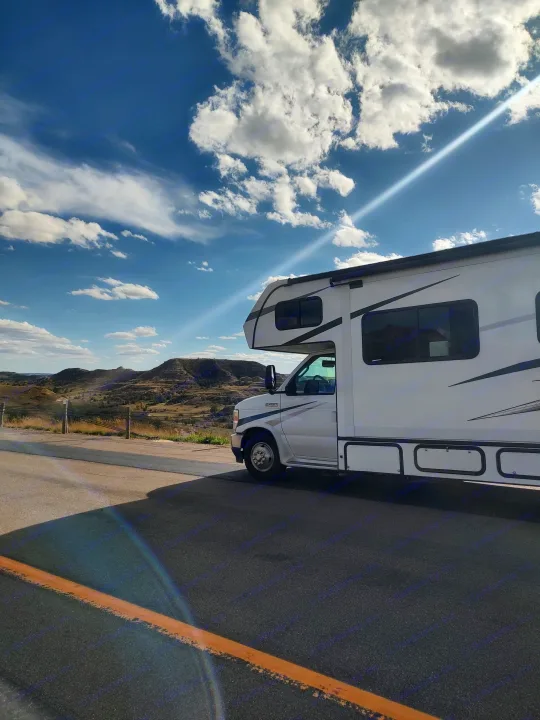 An RV rental parked on a road, offering scenic views of sunlit hills and a blue, cloudy sky with a bright sun ray.. Forest River Sunseeker Classic 2023