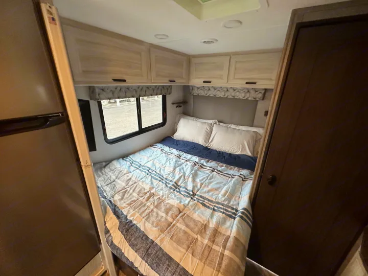 Cozy RV rental bedroom with a striped comforter, built-in storage cabinets, window, and refrigerator for convenience.. Forest River Sunseeker 2022