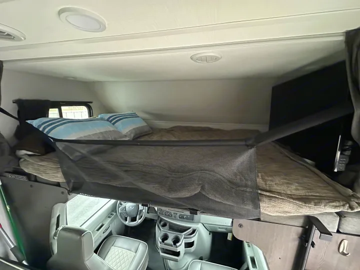 RV rental interior: Overhead view of a cozy bunk bed with safety net above the driver's cab, featuring bedding and a TV.. Forest River Sunseeker 2022