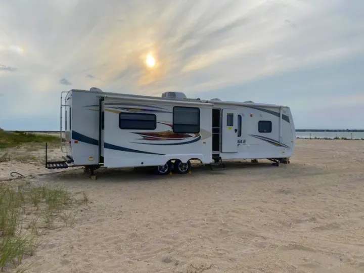 Your perfect RV rental awaits! A spacious RV is set up on a sandy beach by calm waters under a sunlit sky.. Jayco Eagle 2012