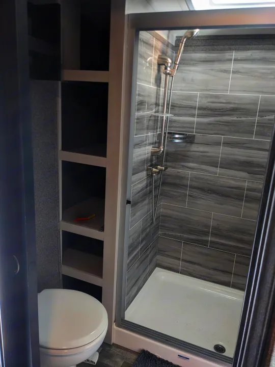 Compact RV rental bathroom with sleek grey-tiled shower, white toilet, and built-in storage shelves. Ideal for travel.. Jayco North Point 2021