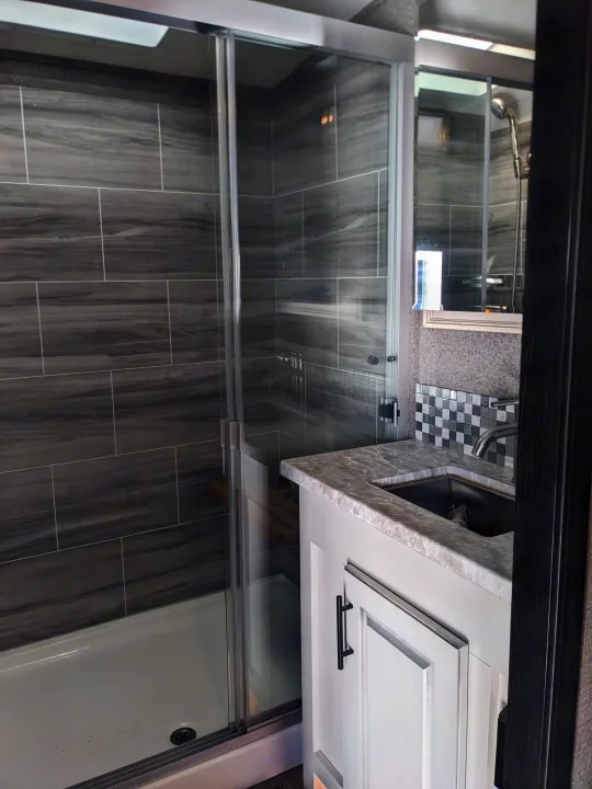 Inside a modern RV rental bathroom: a sleek grey-tiled shower with glass doors, and a vanity with a sink and checkered backsplash.. Jayco North Point 2021