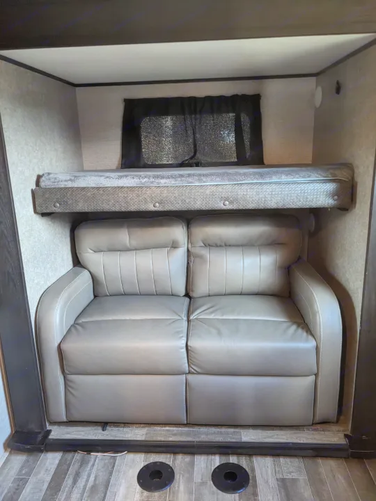 Cozy RV rental interior featuring a gray two-seater sofa beneath an overhead bunk bed and a small window.. Jayco North Point 2021