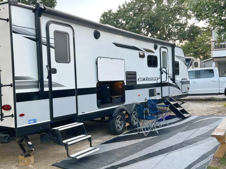 A grey & white Jay Feather RV rental travel trailer is set up at a campsite, featuring an awning, steps, chairs, and rug.. Jayco Jay Feather 2022