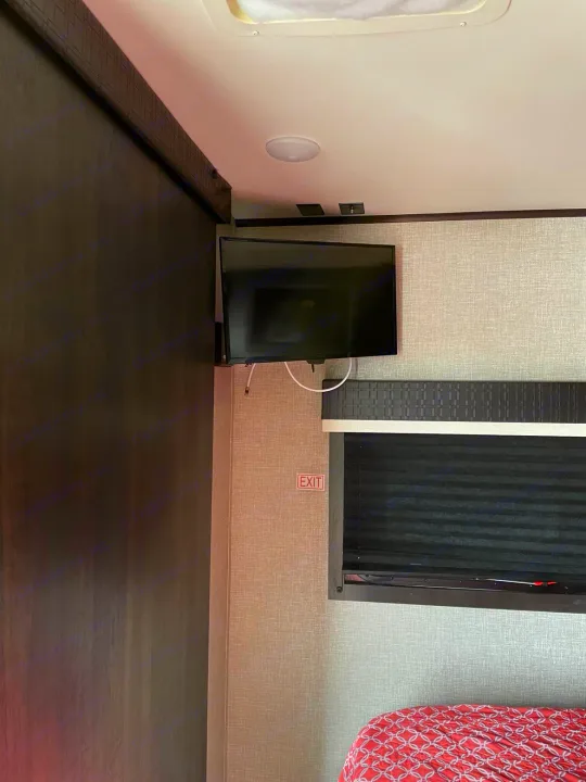 Inside an RV rental bedroom: a mounted TV, dark wood partition, window with blind, and an "EXIT" sign. Cozy RV interior.. Jayco Jay Feather 2022