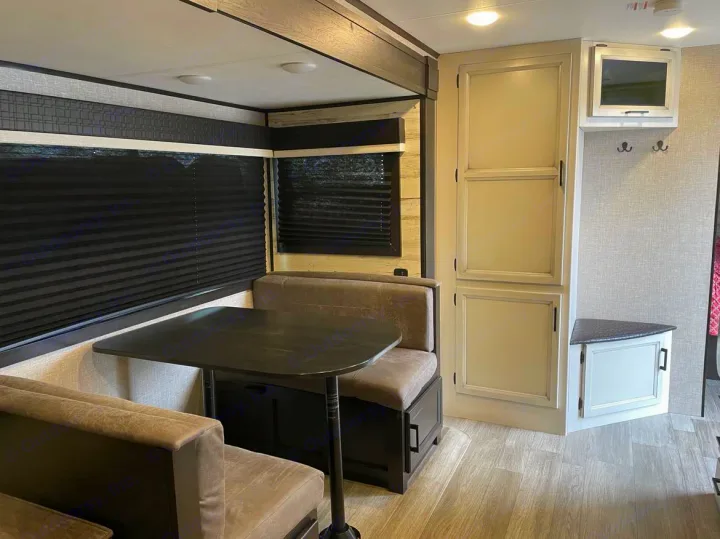 Inside an rv rental: a modern dinette with booth seating, dark table, window blinds, storage cabinets, and a mounted TV.. Jayco Jay Feather 2022