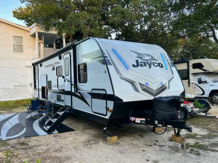Modern Jayco travel trailer RV rental, white with black accents and blue LED lights, parked on gravel with an awning out. Perfect for camping trips.. Jayco Jay Feather 2022