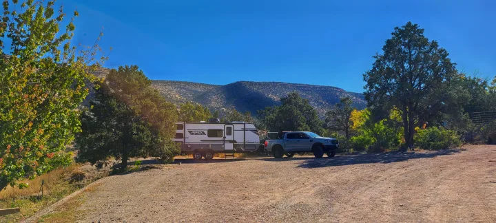 A white rv rental travel trailer and pickup truck are parked on a sunny dirt lot. Trees and mountains frame this scenic outdoor campsite.. Forest River inc Apex 208 BHS 2024