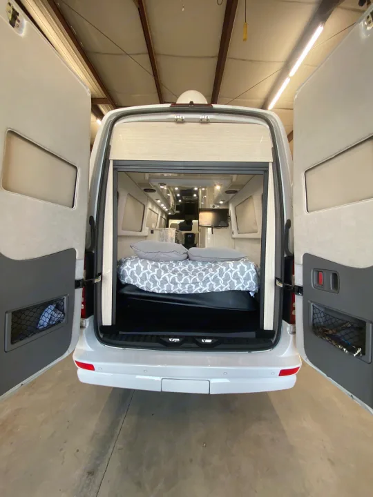 Rear view of an RV rental camper van with its back doors open, showcasing a comfortable bed and interior living area.. Airstream Interstate 2017