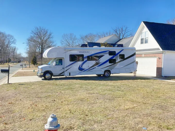 White Chateau RV rental on a driveway in front of a house. A person is partially hidden behind it. Street & trees nearby.. Thor Chateau Victory 2022