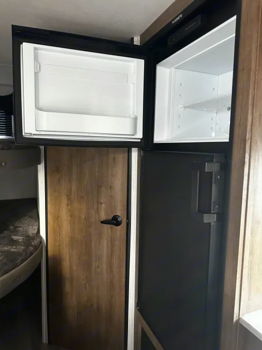 RV rental interior: Open Dometic fridge and freezer. Compact camper kitchen appliances near a wood door and bunk.. Heartland RVs Pioneer 2024