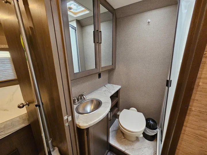 This RV rental bathroom features a metallic sink, marble-look counter, mirrored cabinet, toilet, and textured walls.. Thor Motor Coach Freedom Elite 2021