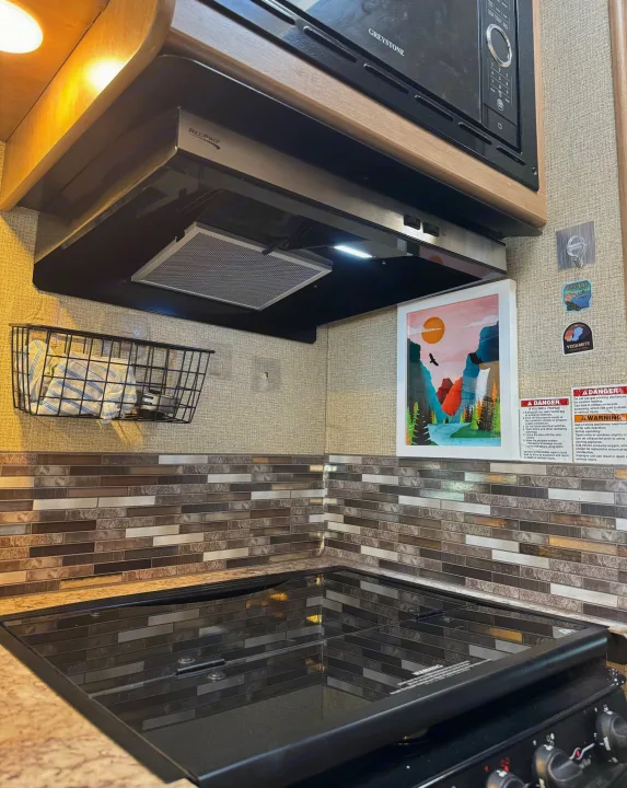 This RV rental kitchen features a compact cooktop, microwave, and range hood, with tiled backsplash, ready for cooking on the go.. Thor Freedom Elite 2019