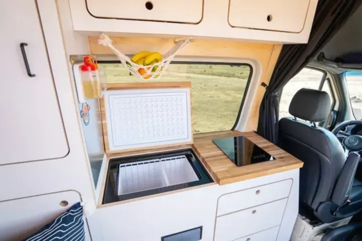 This rv rental features a well-equipped van kitchen with an open fridge, induction cooktop, wood counters, and a fruit hammock.. VanCraft VanCraft Class B 2022