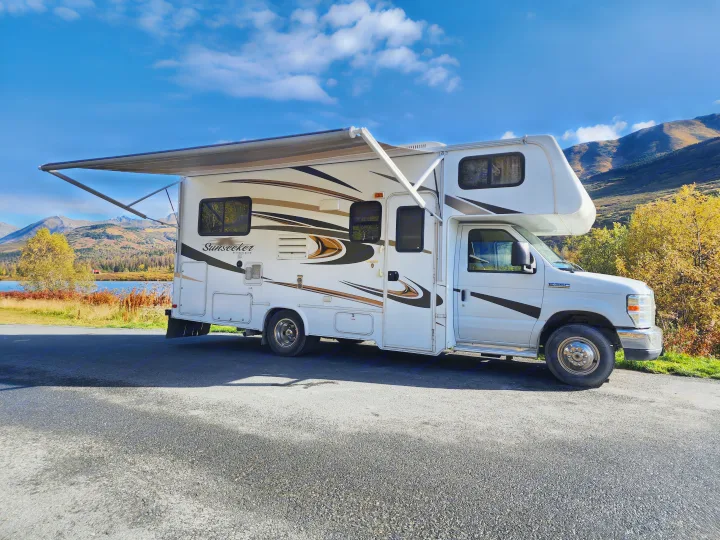 RV rental with awning extended, parked by a scenic lake and autumn mountains under a blue sky. Perfect for travel adventures.. Forest River Sunseeker 2014