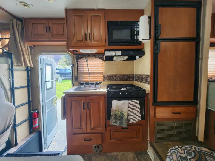 RV rental interior: well-equipped kitchen with microwave, stove, sink, and cabinets. Ladder to bunk, and open door showing outside.. Forest River Sunseeker 2014