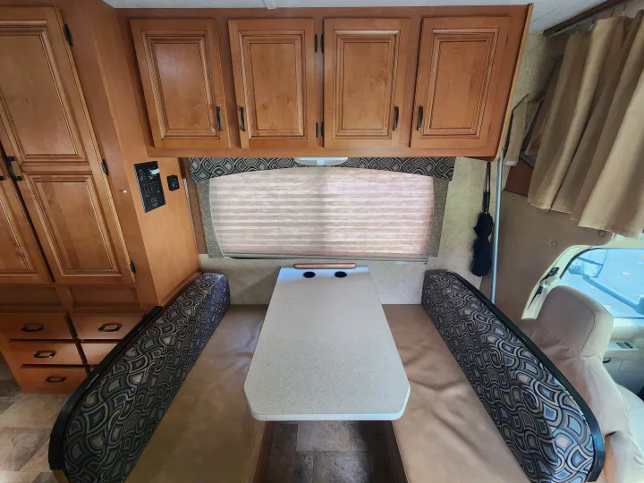 This RV rental interior features a dinette with benches, overhead wood cabinets, and a window blind, perfect for travel comfort.. Forest River Sunseeker 2014