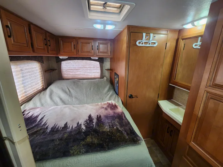 Cozy RV rental interior showing a compact bedroom with a bed, overhead cabinets, and an adjacent bathroom vanity.. Forest River Sunseeker 2014