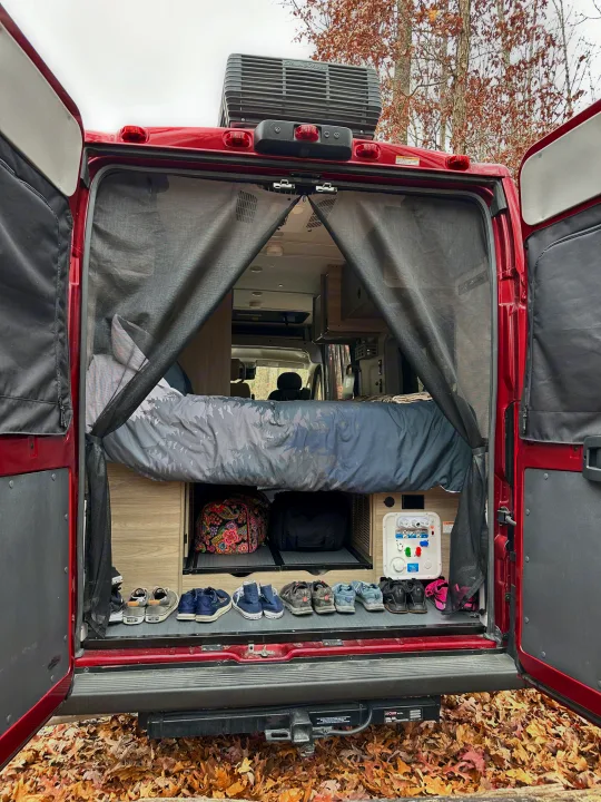 Red RV rental's rear doors open, revealing a cozy bed, shoe storage, and outdoor hookups amidst autumn leaves.. Winnebago Solis 2022