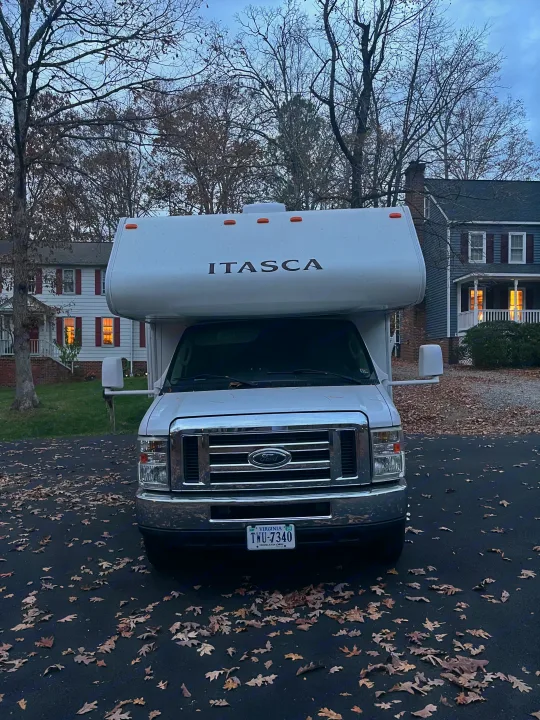 Front view of a white Itasca Class C RV rental parked on a leaf-covered asphalt driveway during autumn, with houses in the background.. Winnebago Winnebago 2014